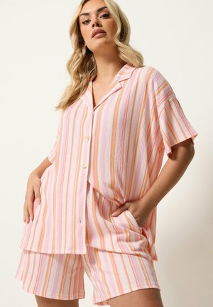 Kvinde iført løs kortærmet skjorte og matchende shorts med lodrette striber i lyserød, orange og hvid, poserer med den ene hånd i lommen.