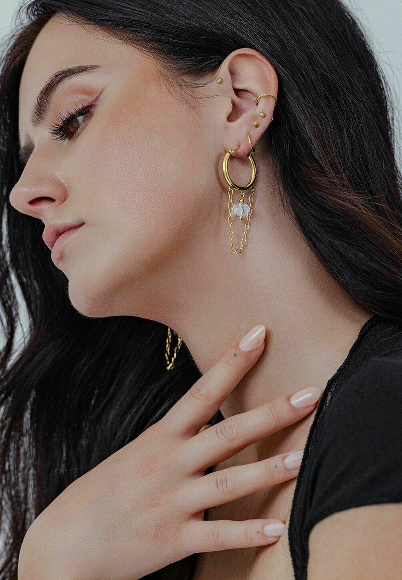 Goldene Creolen mit einem Kristallakzent und Kettendetails, getragen von einem Model mit dunklen Haaren, welches ein Seitenprofil zeigt und eine Hand am Hals hat.