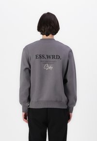 Grå sweatshirt med rund hals, ribbede manchetter og et grafisk print på ryggen med tekst og en signaturdetalje.