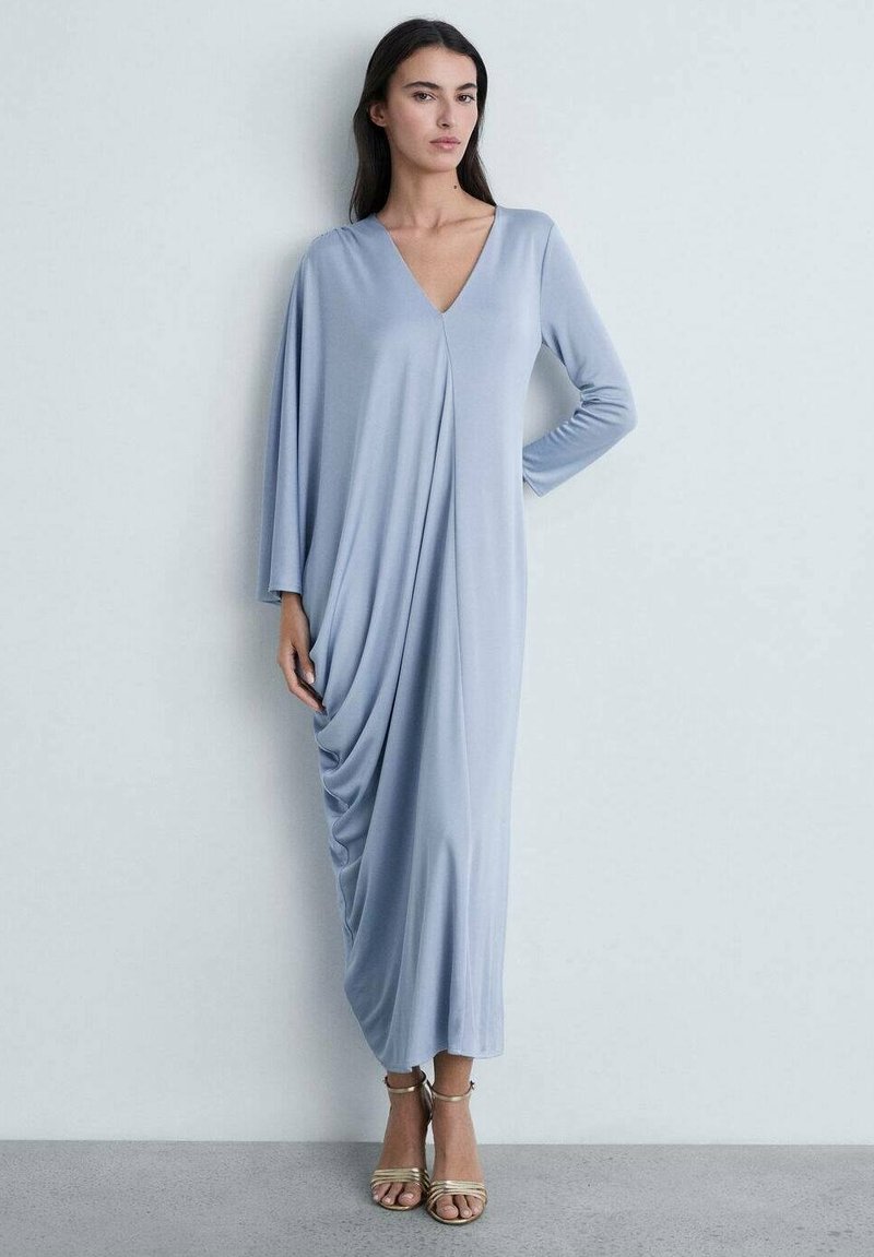 Robe longue drapée en tissu bleu clair avec un profond décolleté en V, des manches longues et un détail froncé sur le côté. Assortie à des sandales dorées à lanières.