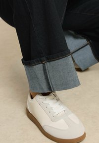 Tummat denim-housut, joissa vaaleat denim-käänteet ja kontrastiksi ommeltu saumavio, yhdistetty valkoisiin tennareihin, joissa on beige pohja.