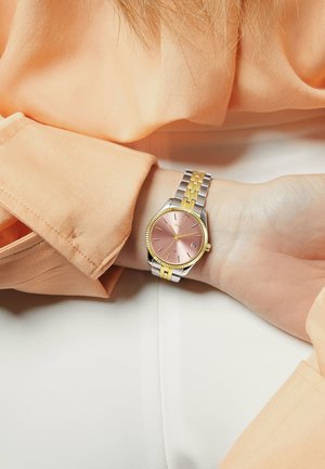 Orologio argento e oro con quadrante rosa, lunetta texturizzata e bracciale in metallo bicolore. Indossato su un polso, sotto una blusa pesca.