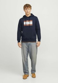 Sweat à capuche bleu marine avec cordons de serrage et poche avant, arborant un logo rectangulaire en blanc et marron. Associé à un jean délavé et des chaussures marron.
