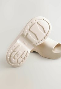 Beige fodtøj med et åbent tådesign. Tekstureret gummisål med skridmønstre. Glat overmateriale med afrundede kanter.