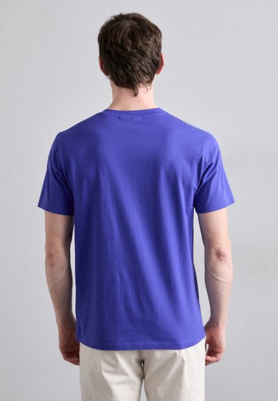 T-shirt en coton violet à manches courtes et col rond, avec une texture lisse et une coupe classique. La vue arrière montre des détails de couture.