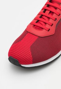 Basket rouge de sport avec une tige en maille et des accents noirs, des détails texturés et une semelle en caoutchouc. Les lacets passent à travers des œillets ronds.