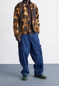 Persona con chaqueta de forro polar marrón y negro con patrón, camisa marrón oscuro, jeans cargo azules sueltos, zapatos verdes y bolso negro de hombro.