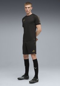 Sort atletisk trøje med guld logo og mønstrede ærmer, parret med shorts med et gult logo. Sorte fodboldsokker og støvler fuldender looket.