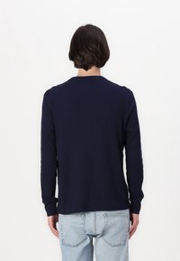 Mörkblå långärmad ribbad tröja med rund halsringning och avslappnad passform, bärs tillsammans med ljusblå jeans, sedd bakifrån.