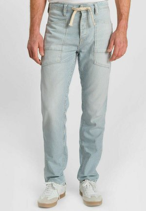 Hombre con pantalones de rayas blancas y azul claro, con grandes bolsillos delanteros y zapatillas blancas, sobre un fondo liso.