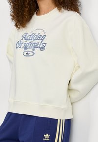 Krämfärgad sweatshirt med ribbade ärmslut och nederkant, med blå "Adidas Originals"-logotyp och en liten trefoilikon på bröstet.