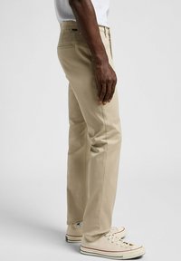Chinos de algodón beige con un corte slim, que cuentan con cremallera, bolsillos laterales y una etiqueta tejida en el bolsillo trasero. Combinados con zapatillas beige.