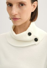 Pull blanc en tricot avec un col montant côtelé et deux boutons noirs sur le col. Texture lisse, design ajusté, style minimaliste.
