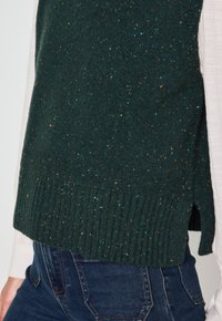 Maglione lavorato a maglia verde scuro con una finitura tessurizzata, caratterizzato da spruzzi d'oro, orlo a coste e spacco laterale, abbinato a jeans blu scuro.