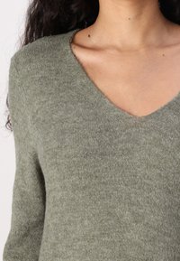 Maglione a V in morbido tessuto verde oliva testurizzato, con maniche lunghe e vestibilità rilassata. I dettagli includono cuciture sottili intorno allo scollo.