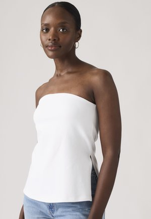 Femme à la peau foncée lisse et aux cheveux attachés portant un haut blanc sans bretelles et un jean bleu clair sur un fond uni.