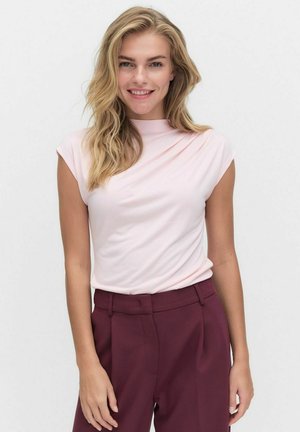 Glimlachende vrouw met lang blond haar, gekleed in een lichtroze mouwloos topje en hoogopgezette donker bordeauxrode broek, staand tegen een witte achtergrond.