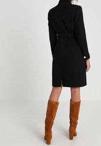 Abrigo de lana negro con cintura ceñida por un cinturón, costuras en los hombros y una abertura en la parte trasera. Combinado con botas de ante marrón hasta la rodilla con tacones de bloque.
