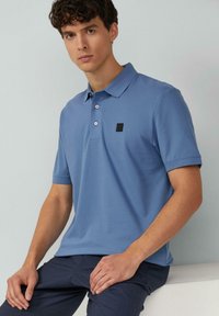 Hellblaues Polohemd aus strukturiertem Baumwollstoff, mit Kragen, drei Knöpfen und einem schwarzen quadratischen Logo-Patch auf der Brust.
