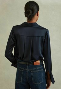 Blusa de seda azul marino con mangas largas, que presenta un pliegue en la parte posterior y puños partidos, combinada con jeans de mezclilla oscuros que muestran una etiqueta de marca de cuero.