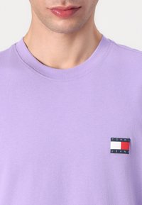 Light purple crewneck t-shirt made of cotton, featuring a rectangular logo patch in navy, red, and white on the left chest area.
