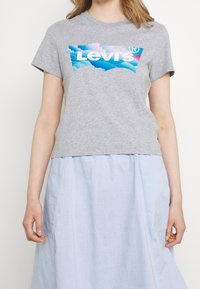 Žena v šedém tričku Levi's s modrým designem oblaku a světle modré sukni, krátké na ramenech a kolenou.