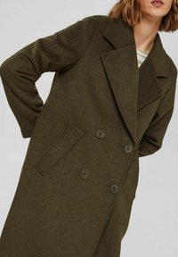 Kobieta ubrana w oliwkowozielony, dwurzędowy płaszcz wełniany z szerokimi klapami i bocznymi kieszeniami, nałożony na koszulę w paski.
