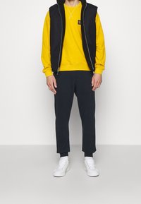 Svart zip-up väst över en gul sweatshirt, i kombination med mörka joggingsbyxor och vita sneakers. Västen har en fleecefoderhuva.