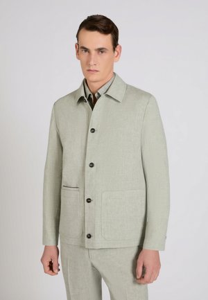 Młody mężczyzna w jasnoszarej zapinanej na guziki kurtce z dwoma przednimi kieszeniami oraz w pasujących spodniach, stojący na jednolitym tle.