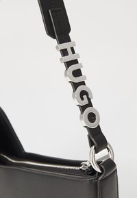 Sac à main en cuir noir avec des accents en métal argenté et le logo "HUGO" embossé sur la sangle. Comprend une ouverture supérieure zippée.