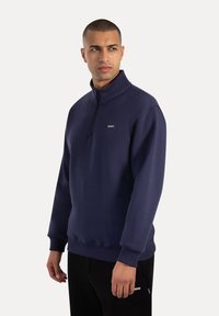 Hombre vestido con un sudadera azul marino de media cremallera y pantalones negros, mirando hacia un lado, aislado sobre un fondo claro.