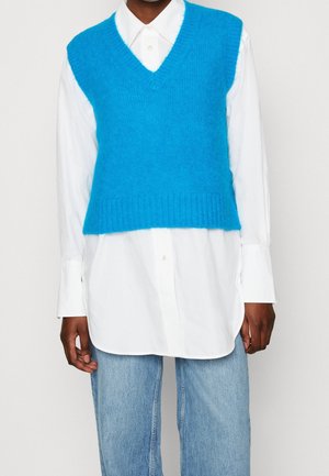 Persoon draagt een felblauwe gebreide mouwloze trui over een lange witte blouse met knopen en lichtblauwe spijkerbroek, staand tegen een effen achtergrond.