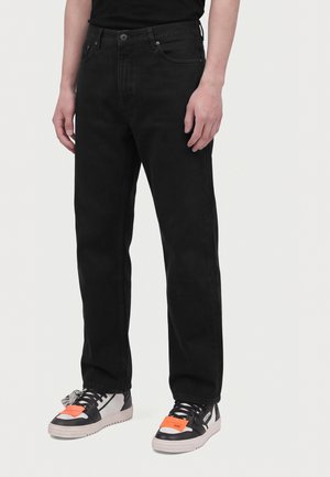 Personne portant un jean noir droit et des baskets noir et blanc avec des étiquettes orange sur un fond blanc uni.