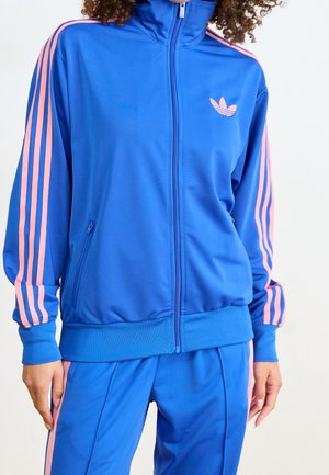 Kvinde iført en blå og pink Adidas træningsjakke og bukser med lynlås foran og logo på brystet, stående mod en ensfarvet baggrund.