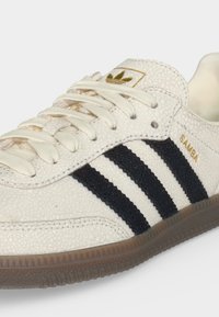 Λευκό sneaker Adidas Samba με μαύρες ρίγες, μπεζ κορδόνια, υφασμάτινο επάνω μέρος και σόλα από καουτσούκ, με χρυσό λογότυπο και λεπτομέρειες επωνυμίας.