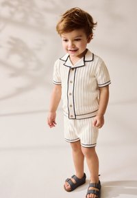 Hvid strikket kortærmet skjorte og shorts med marineblå kant og knapper. Kombineret med mørke sandaler. Lys neutral baggrund.