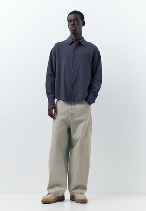 Fiatal férfi áll, kezét a zsebében tartva, sötét gombos ingben, széles világosszürke nadrágban és bézs cipőben egyszerű háttér előtt.