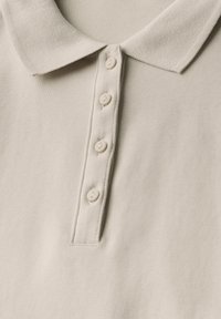 Ljusbeige pikétröja med en strukturerad väv, som har en klassisk krage och fem matchande knappar längs plösen.