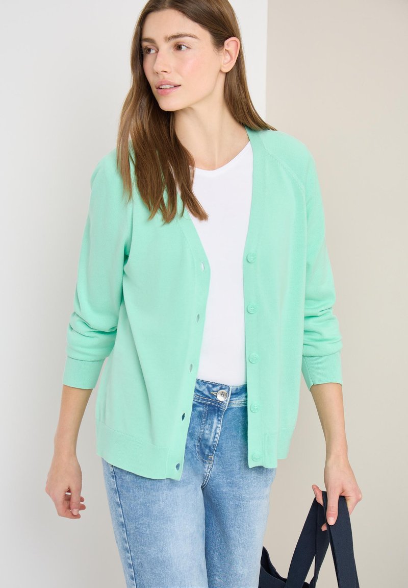Femme portant un cardigan boutonné vert menthe sur une chemise blanche, un jean bleu clair, tenant un sac fourre-tout bleu marine, regardant sur le côté.