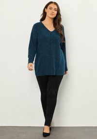 Türkise Strickjacke mit V-Ausschnitt, langen Ärmeln und dezenten Strukturelementen. Kombiniert mit schwarzen Slim-Fit-Hosen und schwarzen Absatzschuhen.