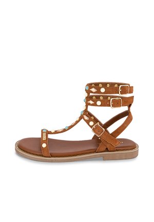 Sandalo in camoscio marrone con multiple fasce regolabili, decorato con borchie turchesi e crema, e suola piatta con bordatura decorativa.