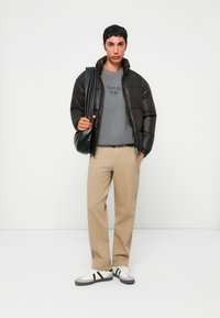 Giubbotto piumino nero, felpa grigia con testo "SURPLUS", pantaloni beige e scarpe da ginnastica bianche con strisce nere. Borsa a spalla nera.