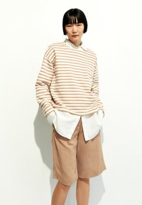 Gestreiftes Langarmshirt in Beige und Weiß, über einem weißen Hemd mit Kragen getragen, kombiniert mit sandfarbenen Shorts. Glatter Stoff mit entspannter Passform.
