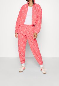 Rosa träningsdräkt med vitt cirkulärt logomönster. Jackan har dragkedja och elastiska muddar; byxorna har resårmidja och muddar.