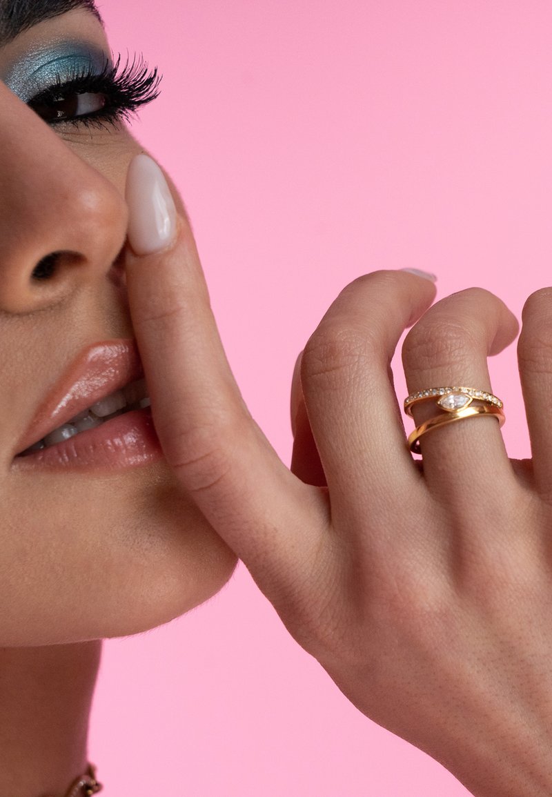 Anelli d'oro con una pietra marquise e una fascia di piccoli dettagli, indossati su un dito. Uno sfondo rosa tenue e ombretto blu esaltano i lineamenti.