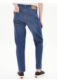 Person som bär blå denimjeans med en lädermärke på midjan, svart- och vit sneakers, står med händerna i bakfickorna.