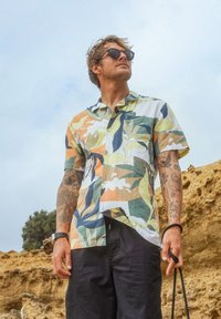 Chemise à manches courtes, à boutons, avec un motif de feuilles multicolores, fabriquée en tissu léger. Portée avec un short sombre et des lunettes de soleil.