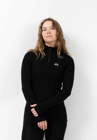 Vrouw met lang golvend haar die een zwart ritsvest met logo draagt, met gekruiste handen voor zich tegen een eenvoudige witte achtergrond.