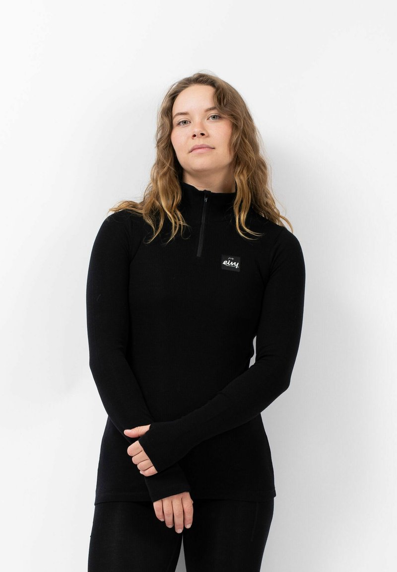 Vrouw met lang golvend haar die een zwart ritsvest met logo draagt, met gekruiste handen voor zich tegen een eenvoudige witte achtergrond.
