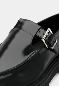 Mocassin en cuir verni noir avec une boucle décorative, des coutures apparentes et un talon légèrement surélevé. Texture lisse et design élégant.